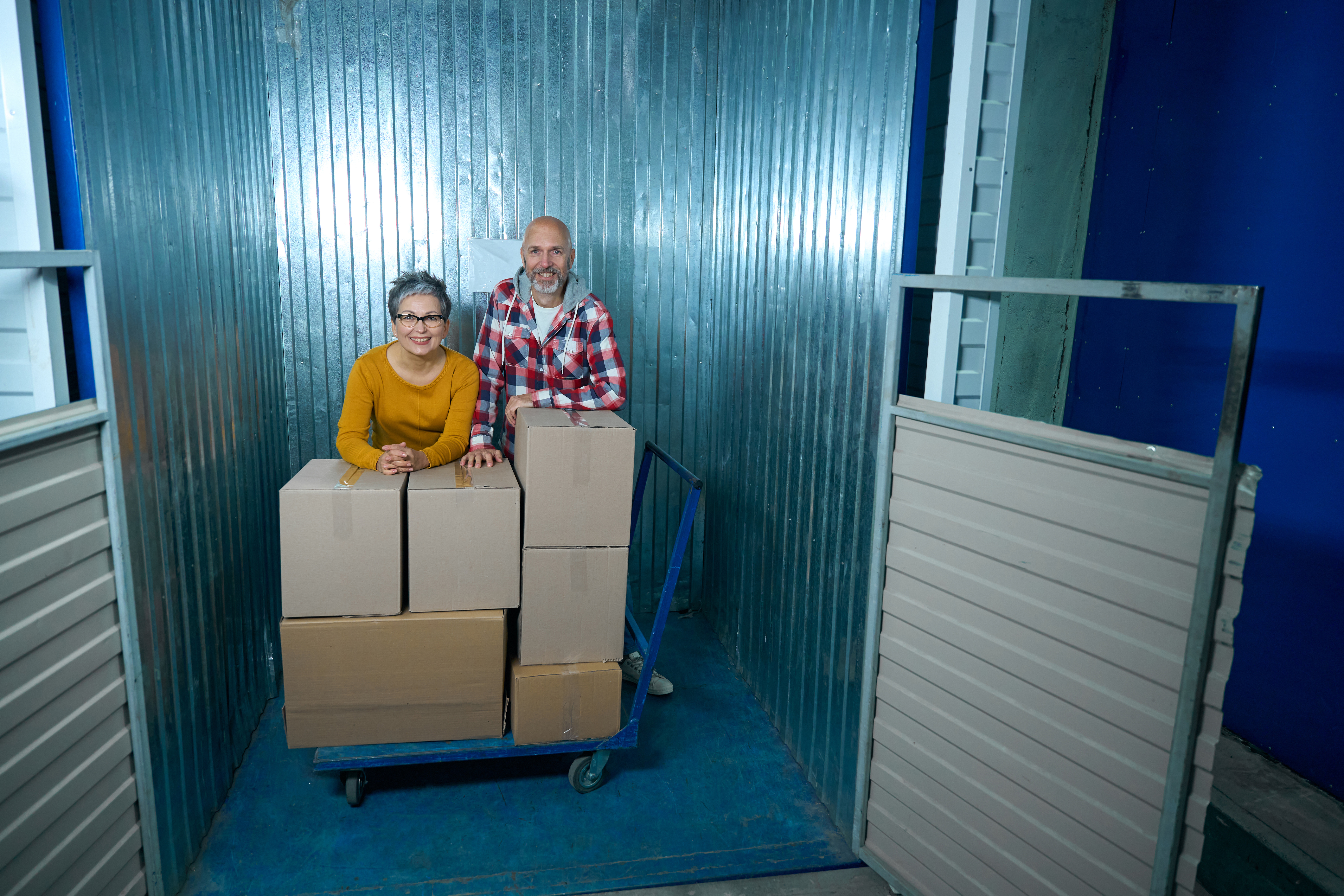 Two people standing with stacked moving boxes inside a storage unit.