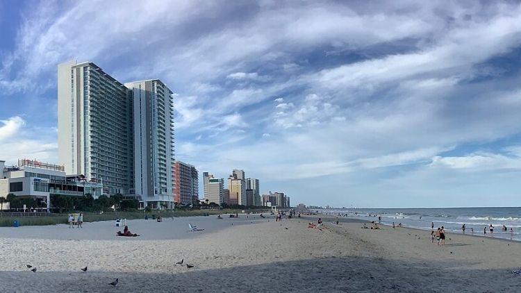 Wide view of Myrtle Beach, South Carolina, showing high-rise hotels and resorts along the sandy shoreline with people relaxing and swimming under a partly cloudy sky.
