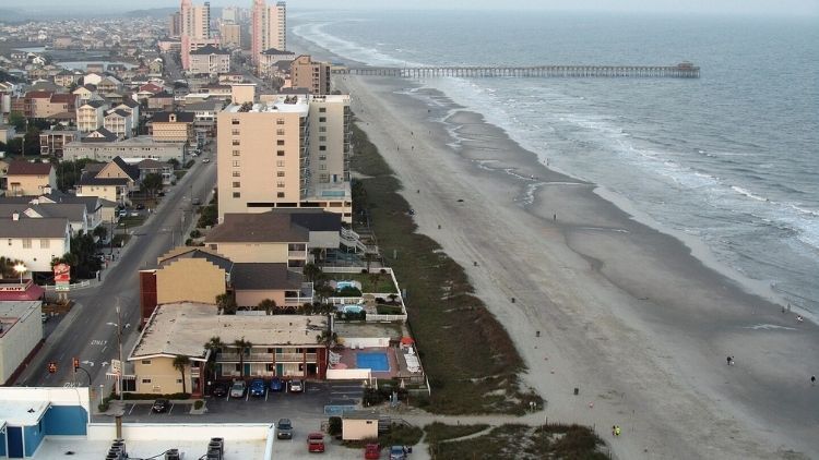 Aerial view of North Myrtle Beach, South Carolina, showing beachfront hotels, residential buildings, a long fishing pier, and the sandy shoreline stretching along the Atlantic Ocean.