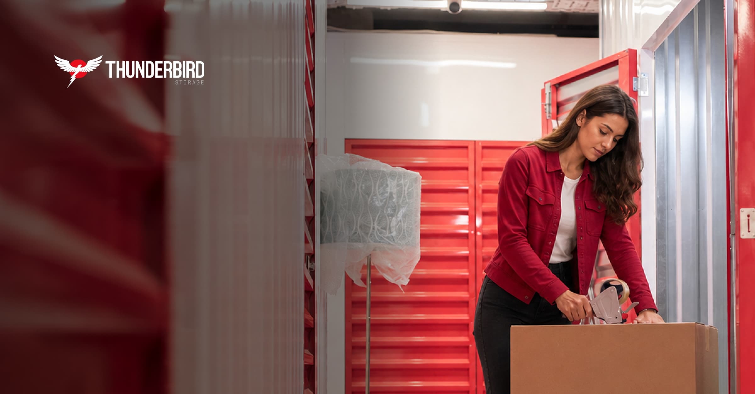 A woman taping a cardboard box to store common household items inside a clean, indoor Thunderbird Storage unit with red doors. A woman taping a cardboard box to store common household items inside a clean, indoor Thunderbird Storage unit with red doors.