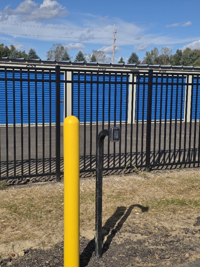 Gate control equipment beside a yellow bollard, with blue storage doors behind the fence.